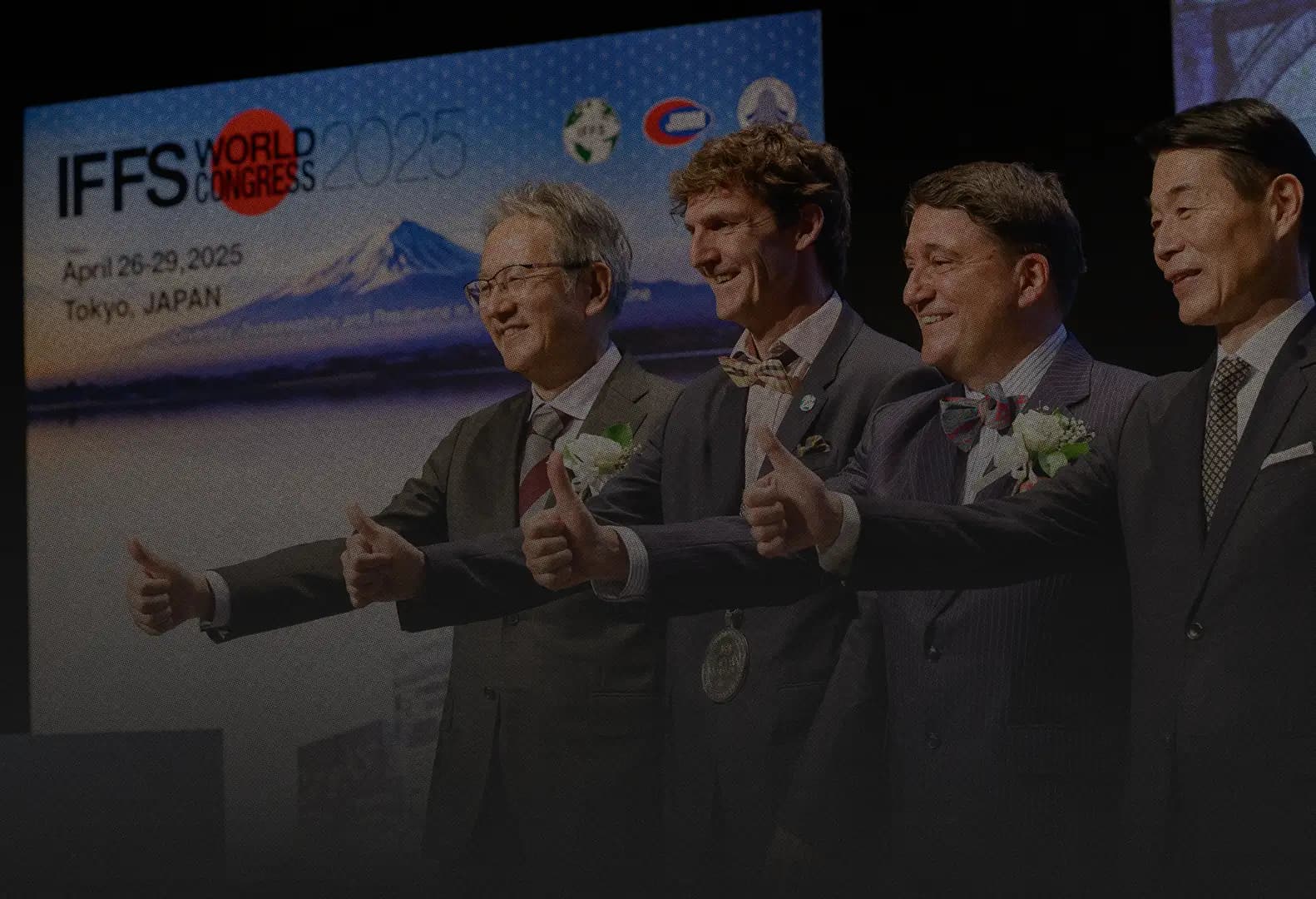 Four men in suits give a thumbs-up on stage at the IFFS World Congress 2025 in Tokyo, in front of a screen showing Mount Fuji and the dates April 26 to 29, 2025.