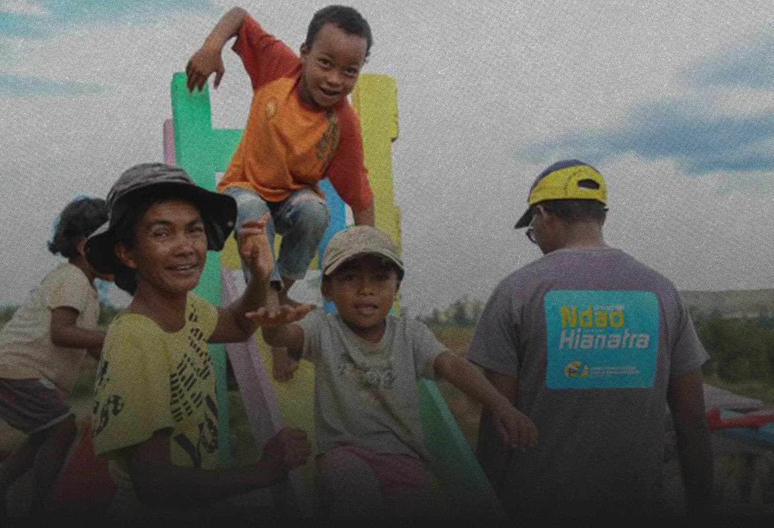 A woman and children smiling on a brightly painted playground slide, beside a man in a “Ndao Hianatra” T-shirt.